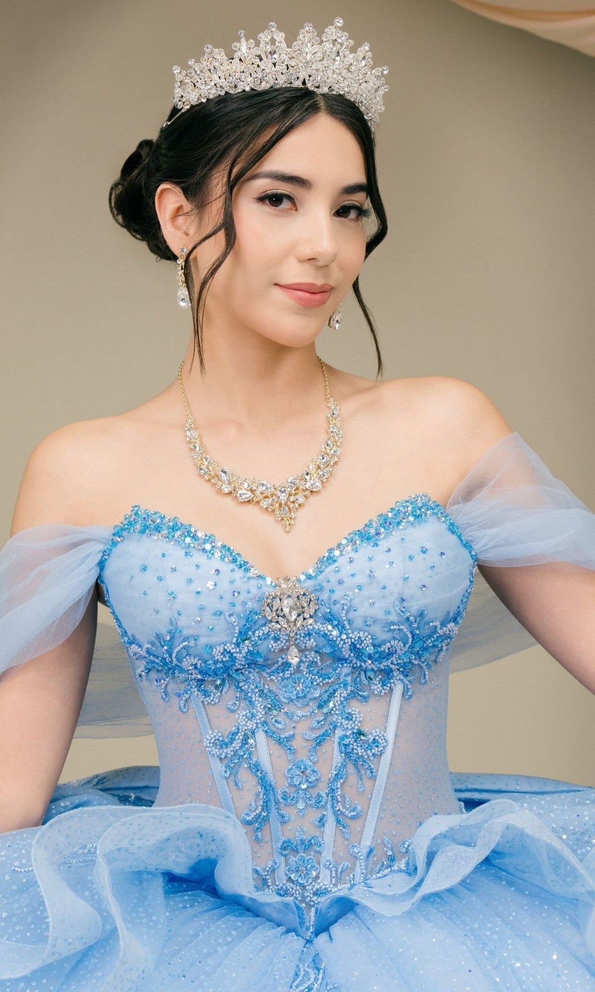 A woman poses gracefully in front of a plain background, wearing the Quinceanera Dress 8520JQ by Cinderella Couture—a dazzling blue ball gown with a corset bodice—paired with a jeweled necklace, earrings, and a sparkling tiara.